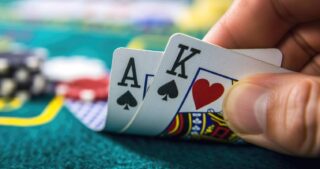 Nahaufnahme einer Hand, die beim Pokerspiel ein Ass und einen König hält; im unscharfen Hintergrund liegen Pokerchips auf grünem Filz.