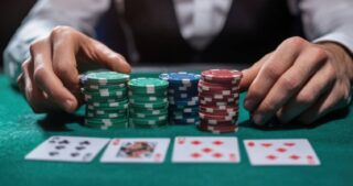 Hände eines Pokerspielers über mehreren Stapeln bunter Chips auf grünem Filz; im Vordergrund liegen vier unscharfe Spielkarten.