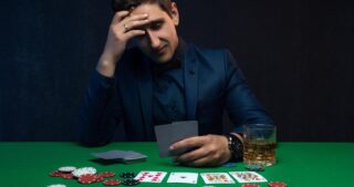 Ein nachdenklicher Mann im Anzug sitzt an einem Pokertisch, hält Karten in der Hand und hat Chips sowie ein Glas Getränk vor sich.